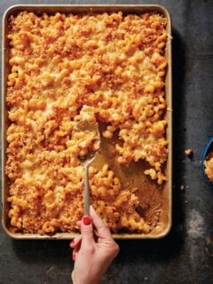 A hand scooping a spoonful of baked macaroni and cheese out of a sheet pan, seen from above.