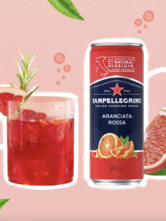 A can of San Pellegrino's Blood Orange flavor can next to a glass of the drink on a designed pink background with blood oranges.