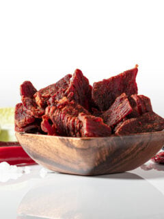 A close up product photo of Tajin flavored beef jerky in a wooden bowl with ingredients on the counter behind it.