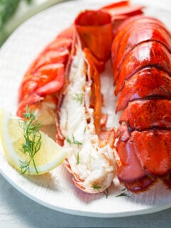 Cooked lobster tails on a white plate with lemon and dill