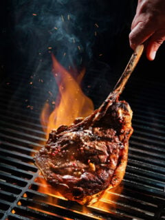 A Tomahawk steak being grilled over fire and being held by the long bone