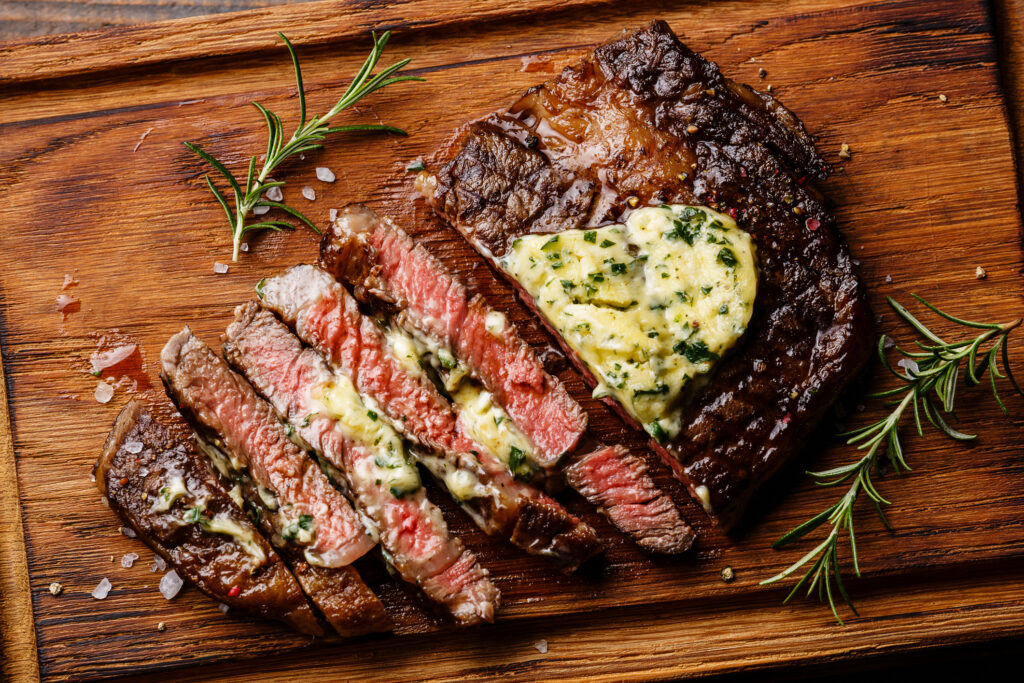 A freshly cooked ribeye steak partially cut into slices on a cutting board and topped with herby butter.