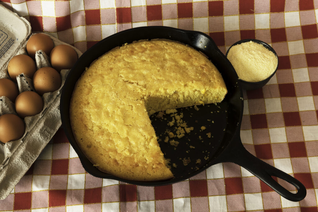 Homemade cornbread in a cast iron skilet, with eggs and a container of corn flour arranged on the side.