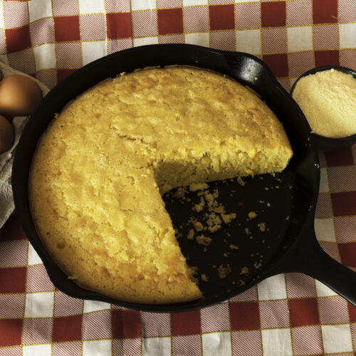 Homemade cornbread in a cast iron skilet, with eggs and a container of corn flour arranged on the side.
