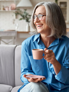 Happy relaxed grey-haired woman drinking coffee relaxing on sofa at home.