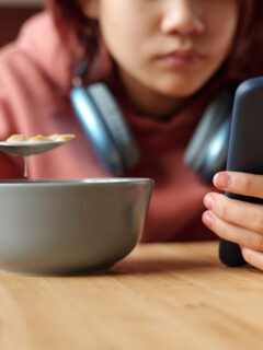 Hand of teenage girl with mobile phone having cornflakes with milk from bowl while sitting by table in front of camera and reading messages