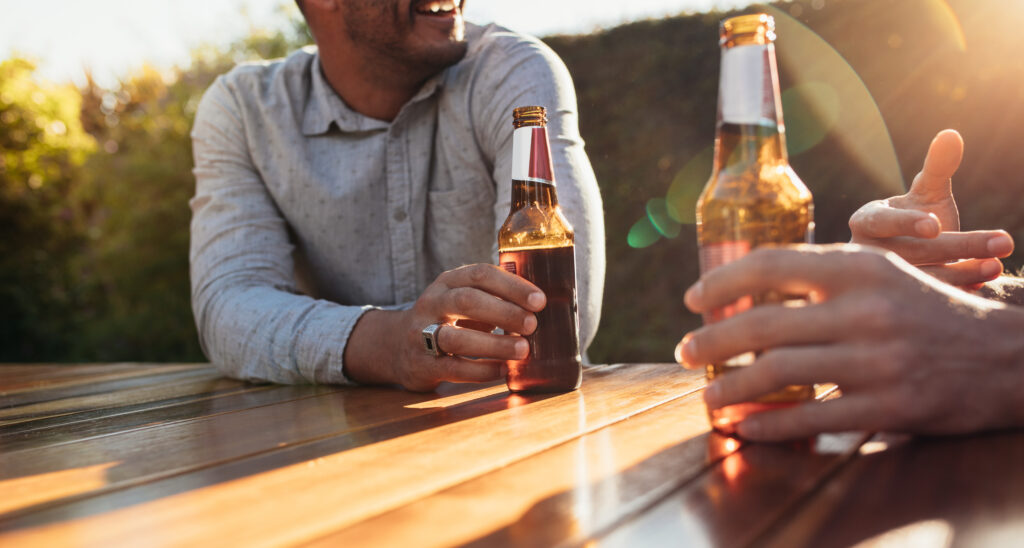 Couple sitting outdoors at wooden table having beers and talking. Focus on couple hands with beer bottles during a outdoor party.