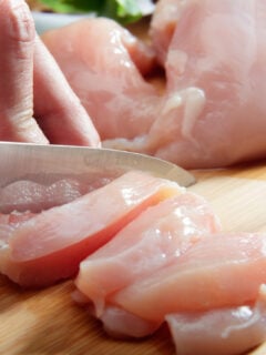 Man's hand cutting raw chicken breast with a knife on a cutting board.