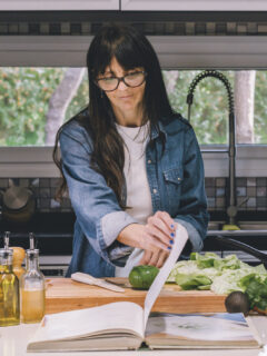 Woman reading recipe from a cookbook while preparing food in the kitchen.