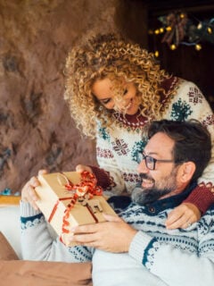 Happy couple enjoy christmas sharing gifts leisure activity at home. Xmas decorations and tree in background. Man receiving a surprise box present from his wife.