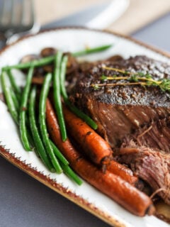Pot roast being served on a white oval platter green beans and carrots. Gray tablecloth.