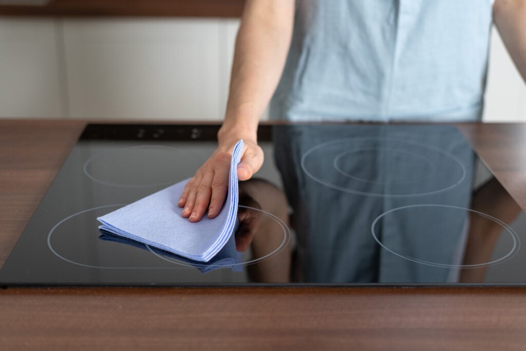 Housework and cleaning service concept. Cropped view of woman in apron wiping induction stove with ceramic surface, standing on modern kitchen and using cloth rag
