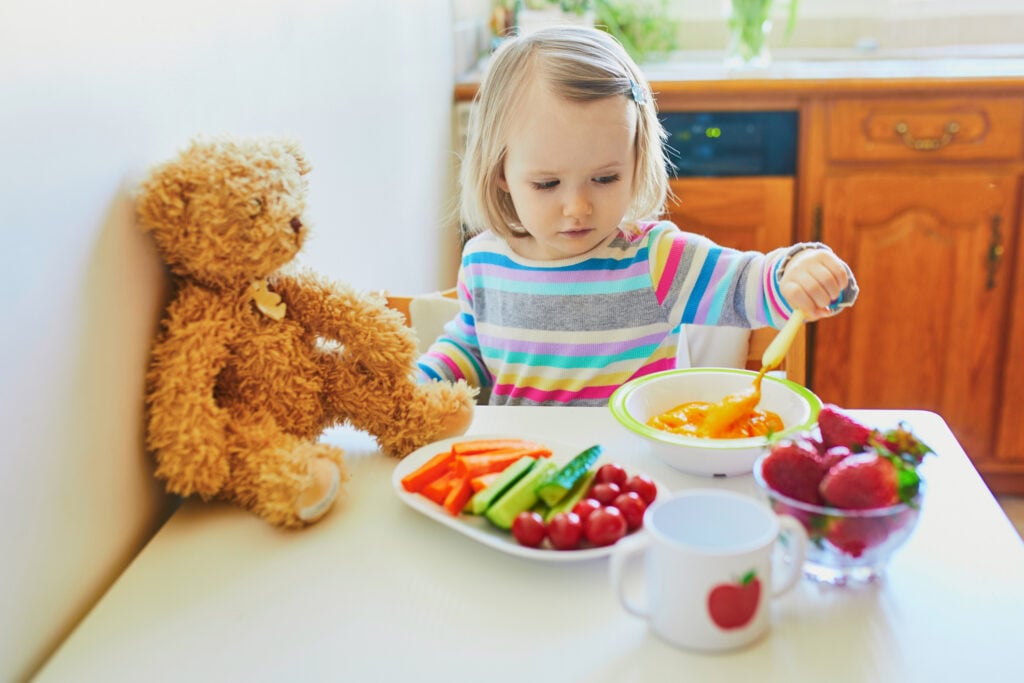 Adorable toddler girl eating fresh fruits and vegetables for lunch. Child feeding teddy bear in the kitchen. Delicious healthy food for kids